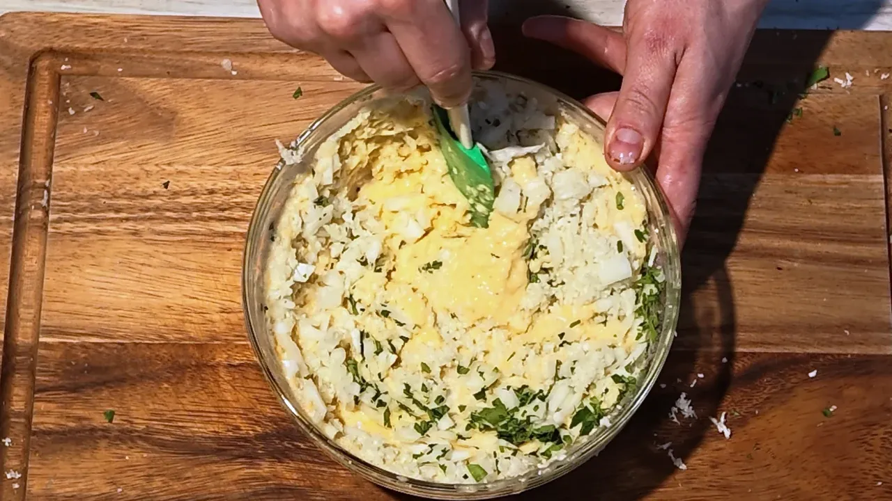 Overhead view of grated cauliflower, onion and parsley being stirred into the batter with a green spatula