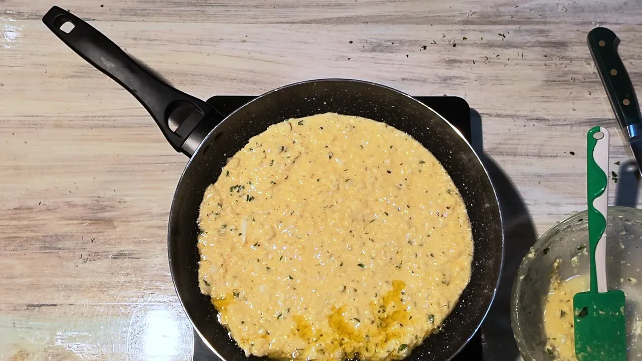 Overhead view of the batter spread into one round in a black non-stick pan on an induction hob