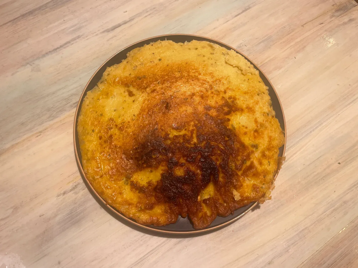 Overhead view of a round golden-brown cauliflower chickpea omelette, darker in the middle, on a dark plate on a pale wooden table