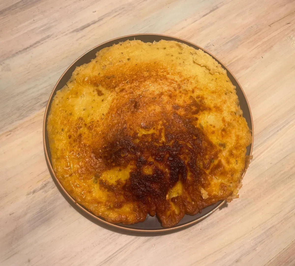 Overhead view of a round golden-brown cauliflower chickpea omelette, darker in the middle, on a dark plate on a pale wooden table