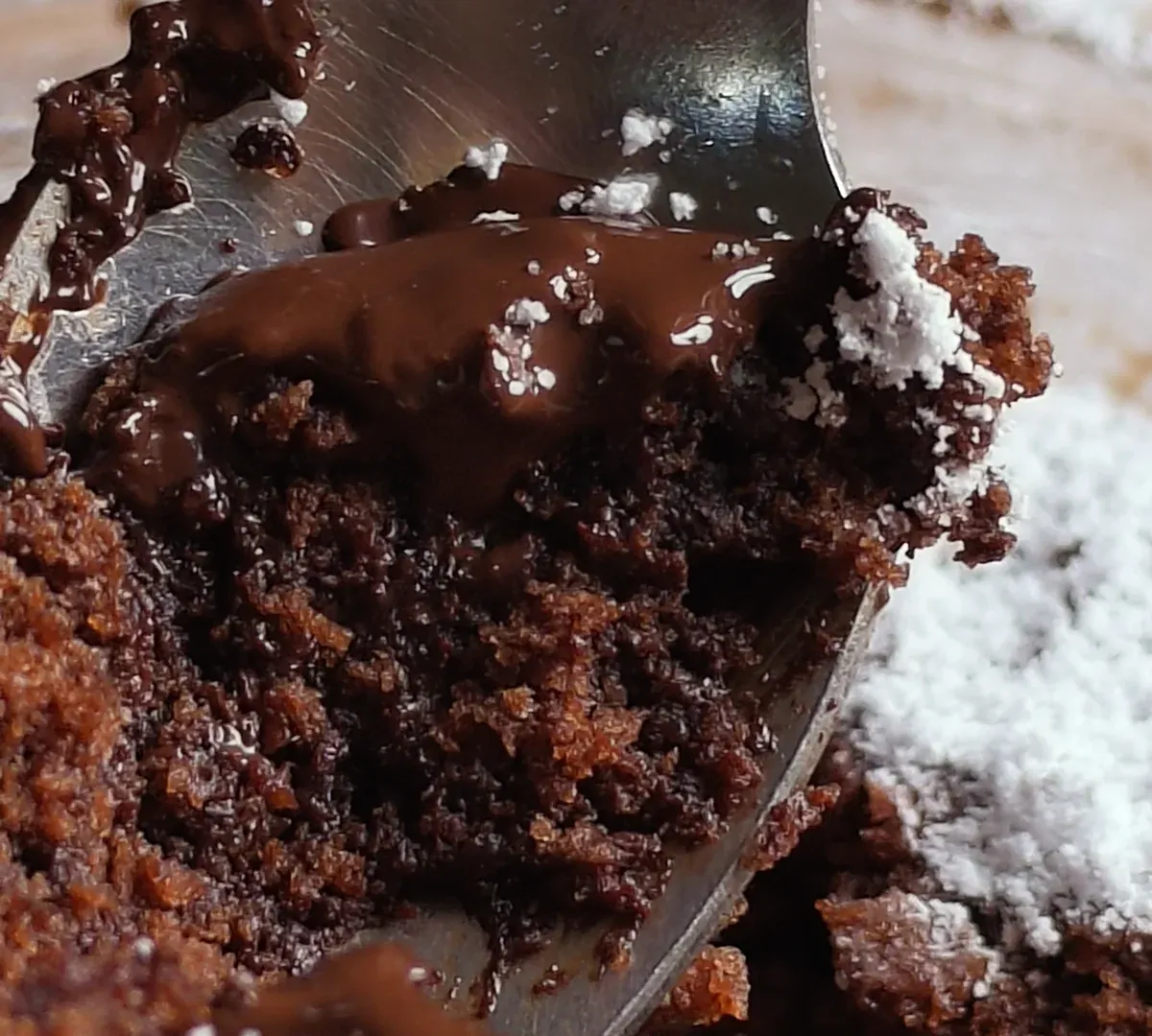 Close-up of a teaspoon lifting soft chocolate cake and molten dark chocolate out of a bowl dusted with icing sugar
