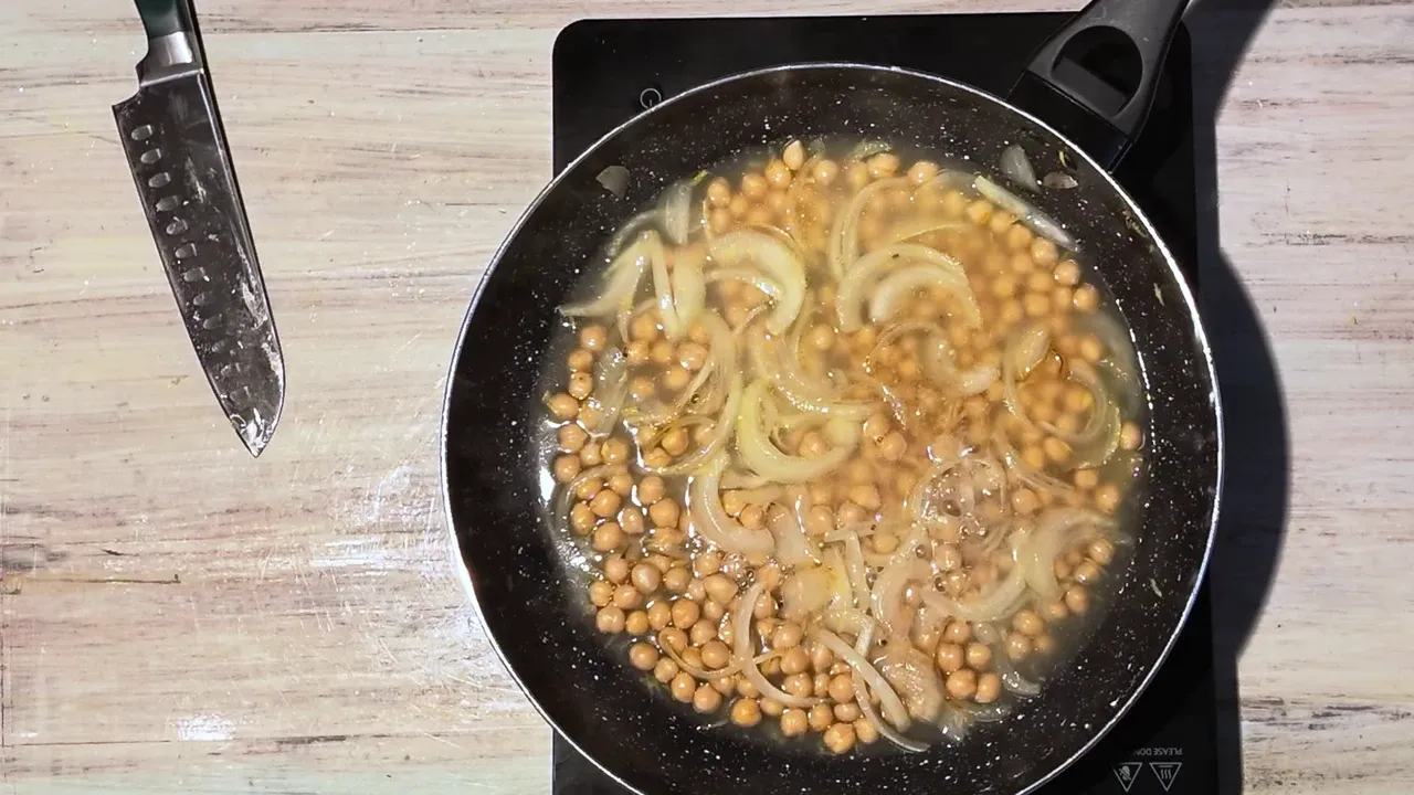 Overhead view of chickpeas and onion covered with broth in a black pan beside a chef's knife