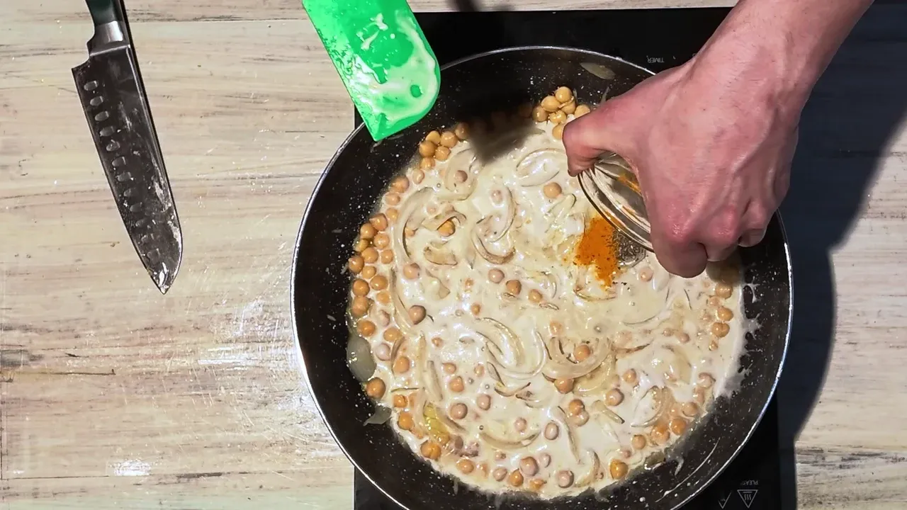 A hand tipping turmeric and black pepper into chickpeas just stirred with coconut milk