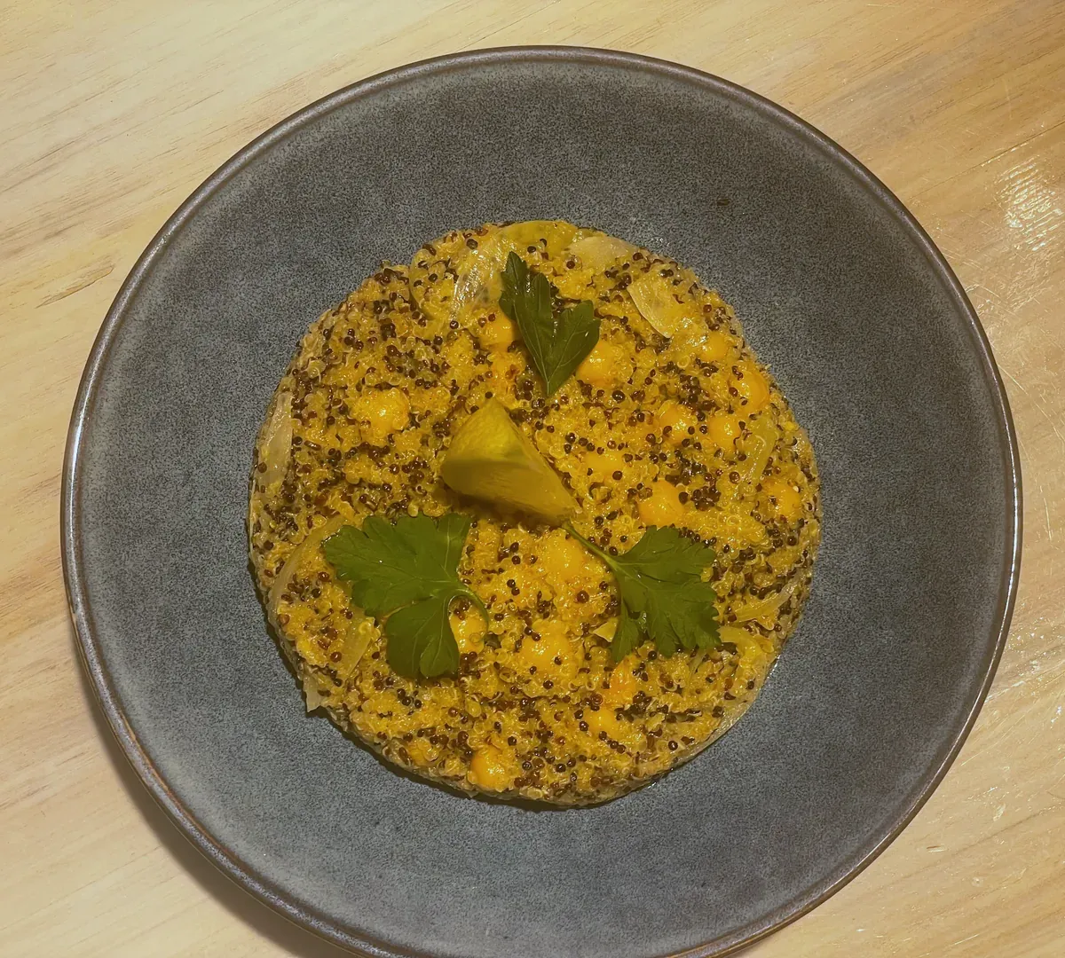 Overhead view of a grey ceramic plate on a wooden table holding a round of golden quinoa, chickpeas and soft onion slices, topped with three parsley leaves and a lemon wedge