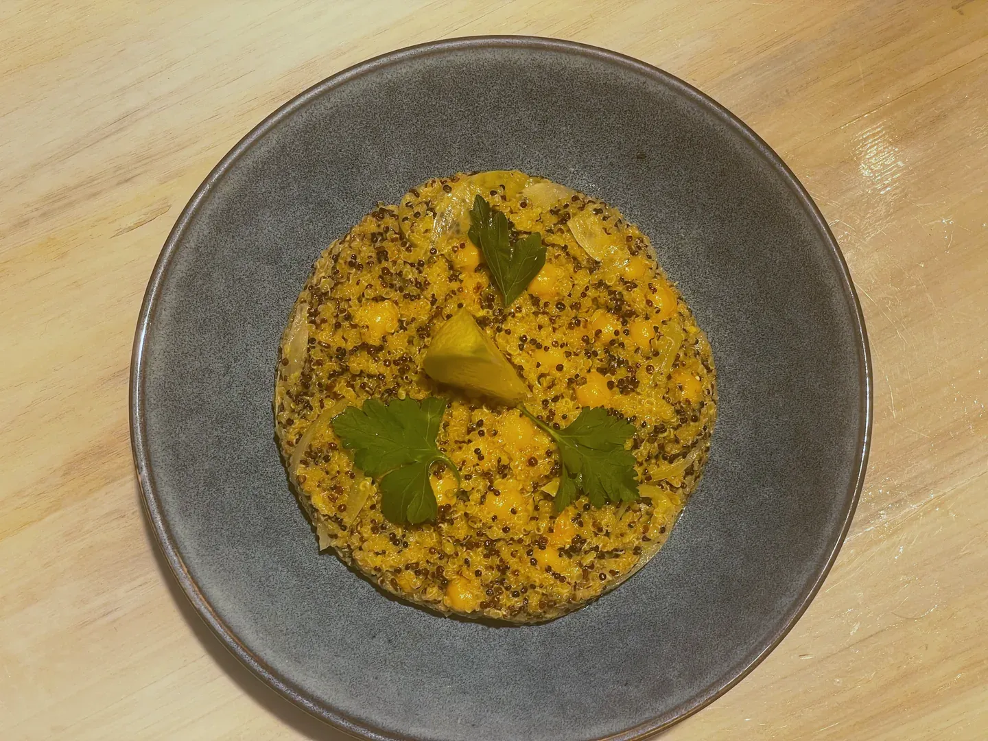 Overhead view of a grey ceramic plate on a wooden table holding a round of golden quinoa, chickpeas and soft onion slices, topped with three parsley leaves and a lemon wedge