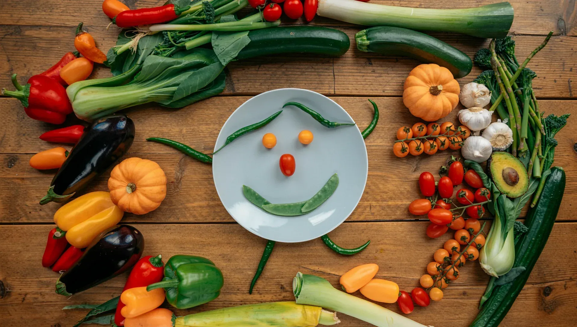 A plate with a smiley face made from vegetables, surrounded by fresh colorful produce