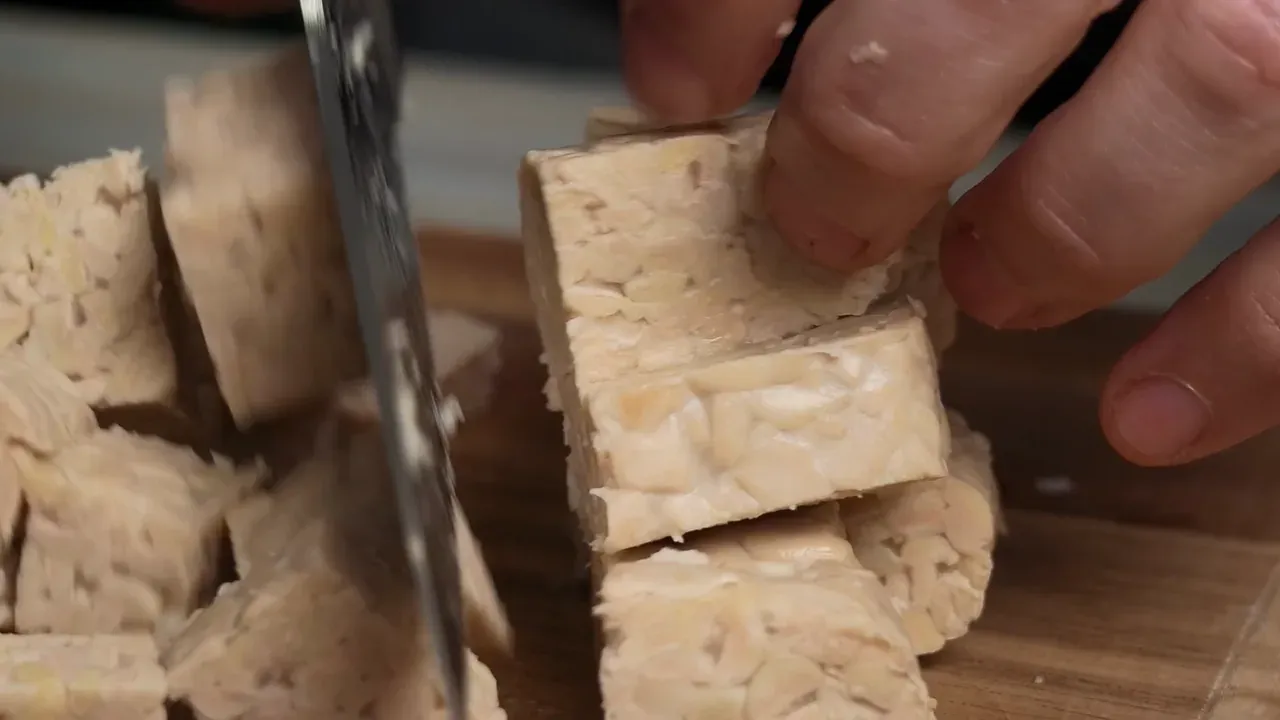 A knife cutting a strip of tempeh into cubes on a wooden board