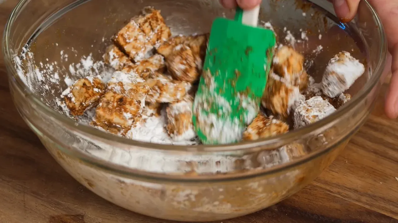 Spiced tempeh cubes dusted with white cornstarch, turned with a green spatula in a glass bowl