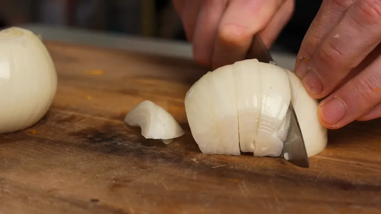 A knife cutting half a peeled onion into wedges on a wooden board