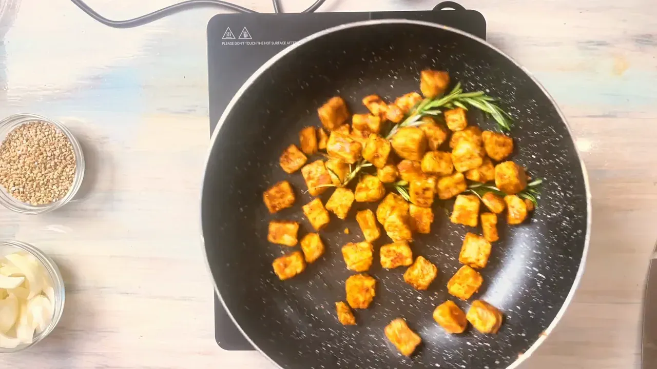 Golden tempeh cubes with rosemary sprigs in a black non-stick pan, seen from above