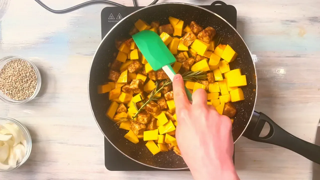 Pumpkin cubes and golden tempeh stirred with a green spatula in the pan, seen from above