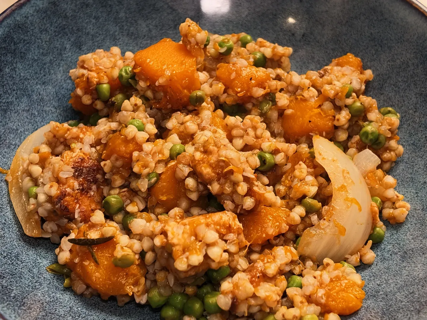 A blue ceramic bowl of buckwheat with orange pumpkin cubes, green peas, browned tempeh and soft onion petals