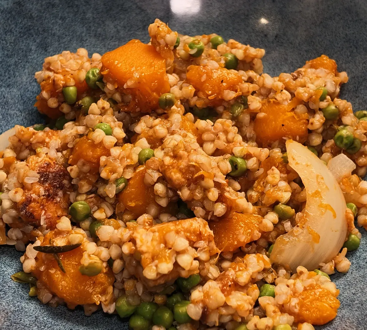 A blue ceramic bowl of buckwheat with orange pumpkin cubes, green peas, browned tempeh and soft onion petals