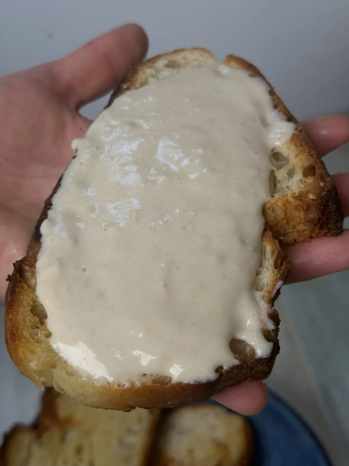 A slice of toasted crusty bread covered with a thick layer of creamy, glossy vegan cheese, held in a hand above a plate of toast