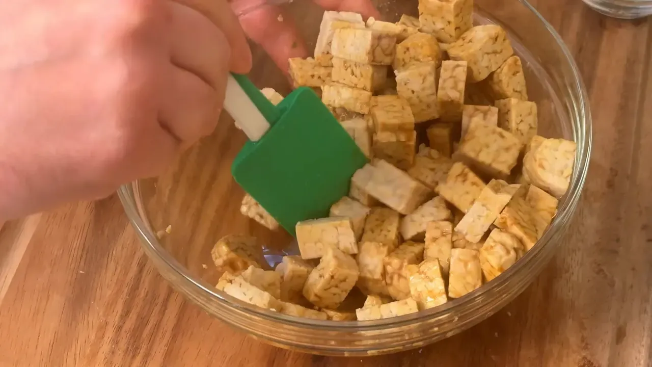 Tempeh cubes being tossed in soy sauce and olive oil with a green spatula in a glass bowl