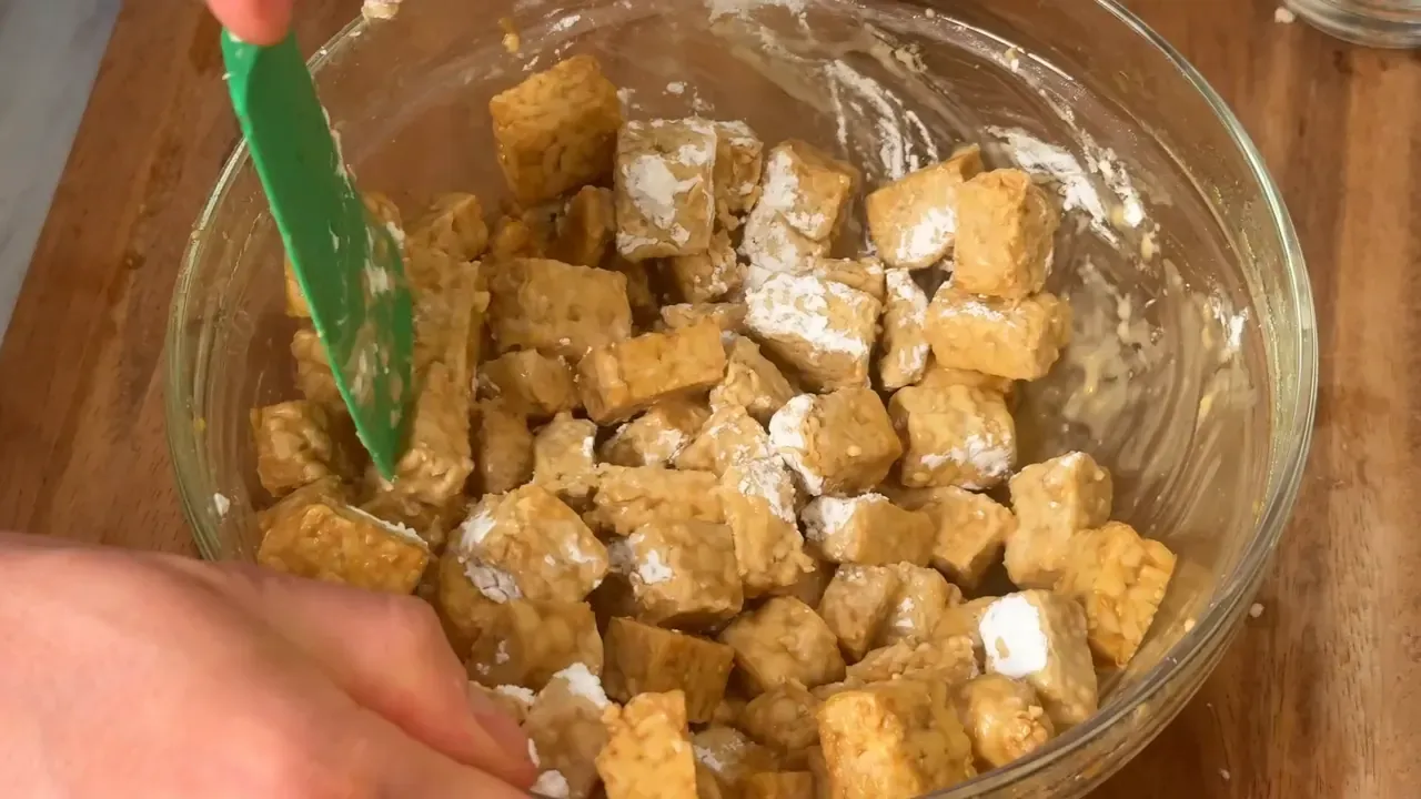 Marinated tempeh cubes dusted with white cornstarch in a glass bowl, turned with a green spatula