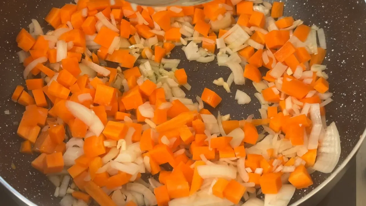 Diced carrot and chopped onion sautéing in olive oil in a dark pan