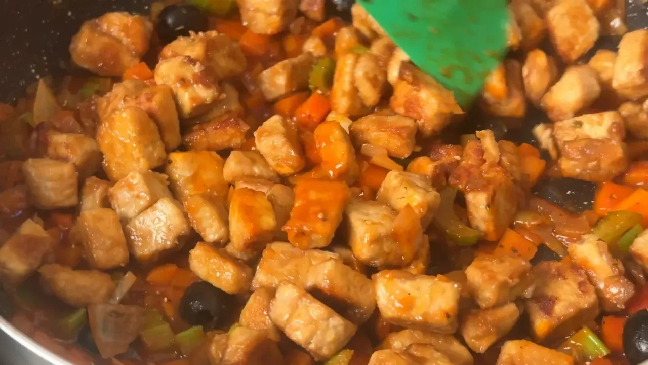 Browned tempeh cubes being stirred into the tomato and vegetable sauce with a green spatula