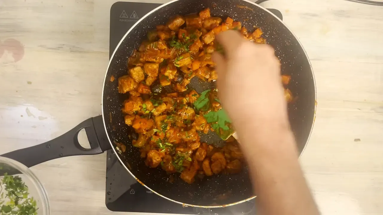 A hand scattering chopped fresh parsley over the tempeh stew in the pan, seen from above