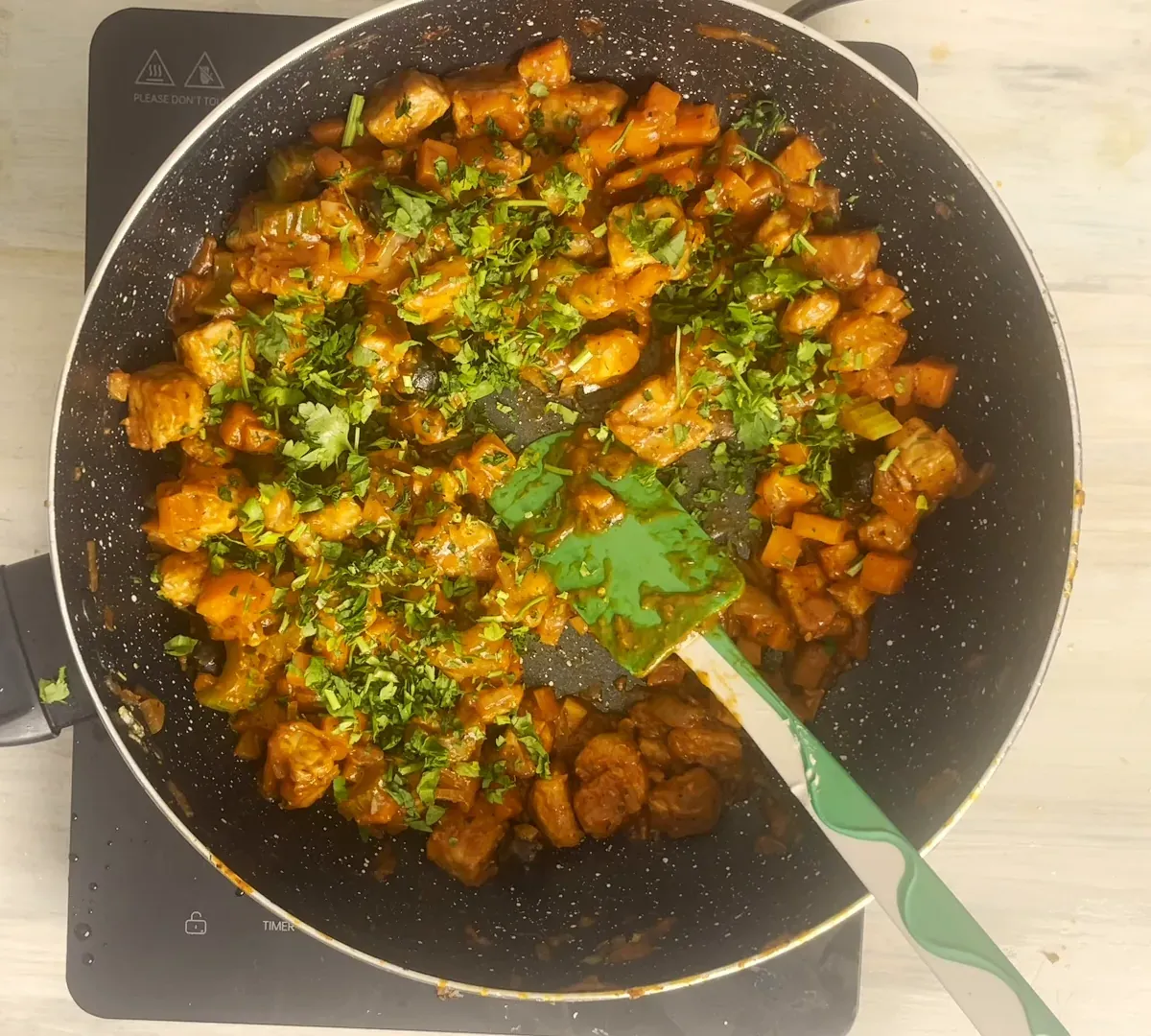 Overhead view of tempeh cacciatora in a dark speckled pan: browned tempeh cubes and diced carrot in tomato sauce, scattered with chopped fresh parsley, a green spatula resting in the pan