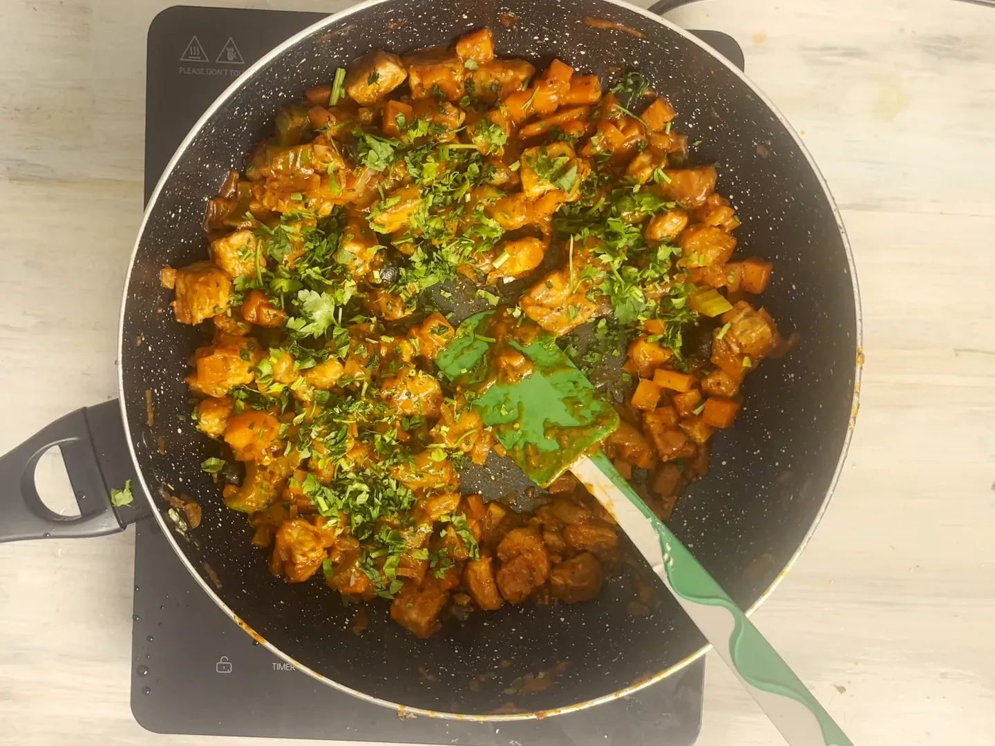 Overhead view of tempeh cacciatora in a dark speckled pan: browned tempeh cubes and diced carrot in tomato sauce, scattered with chopped fresh parsley, a green spatula resting in the pan
