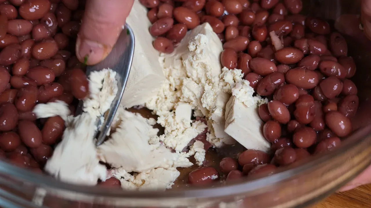 A fork breaking a block of tofu into red kidney beans in a glass bowl