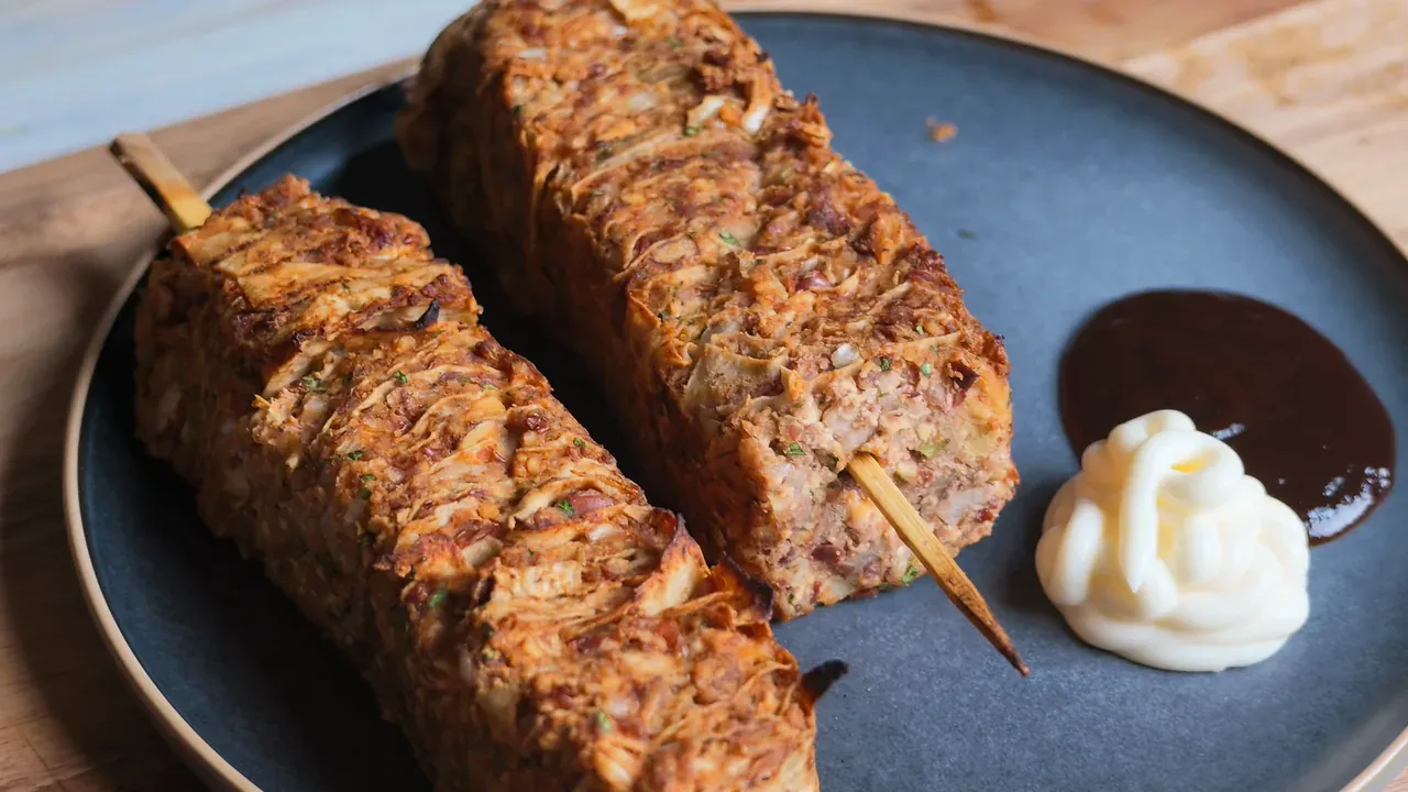Two baked skewers on a dark plate with vegan mayonnaise and dark BBQ sauce