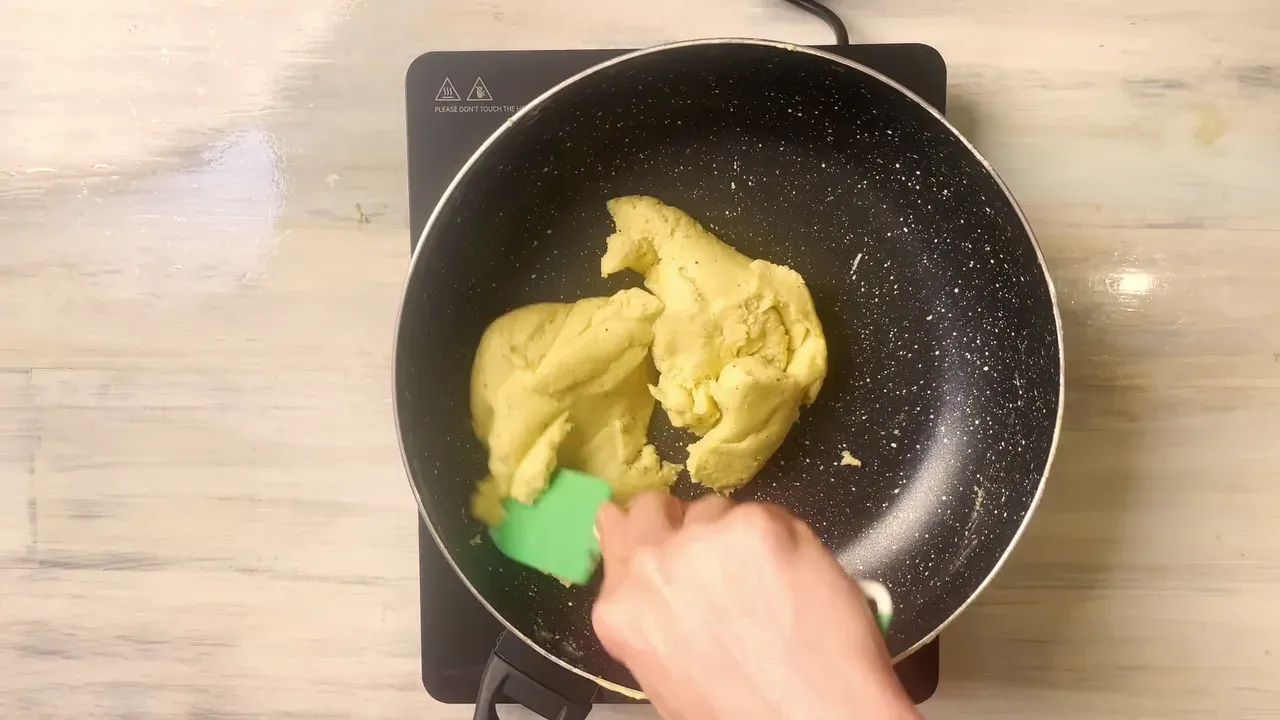 Una mano raccoglie con una spatola verde l'impasto cotto e compatto in una padella nera su un piano a induzione, visto dall'alto