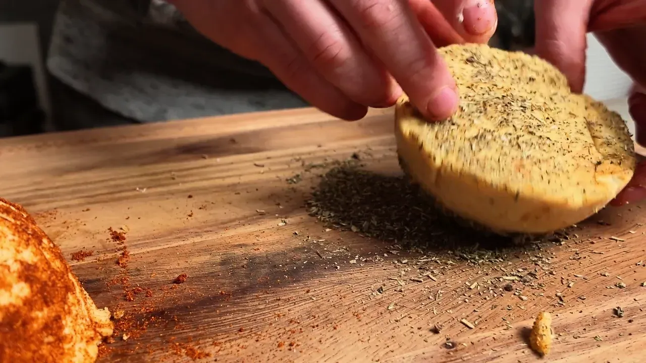 Mani che passano una caciotta nell'origano secco su un tagliere di legno, accanto a una caciotta ricoperta di paprica