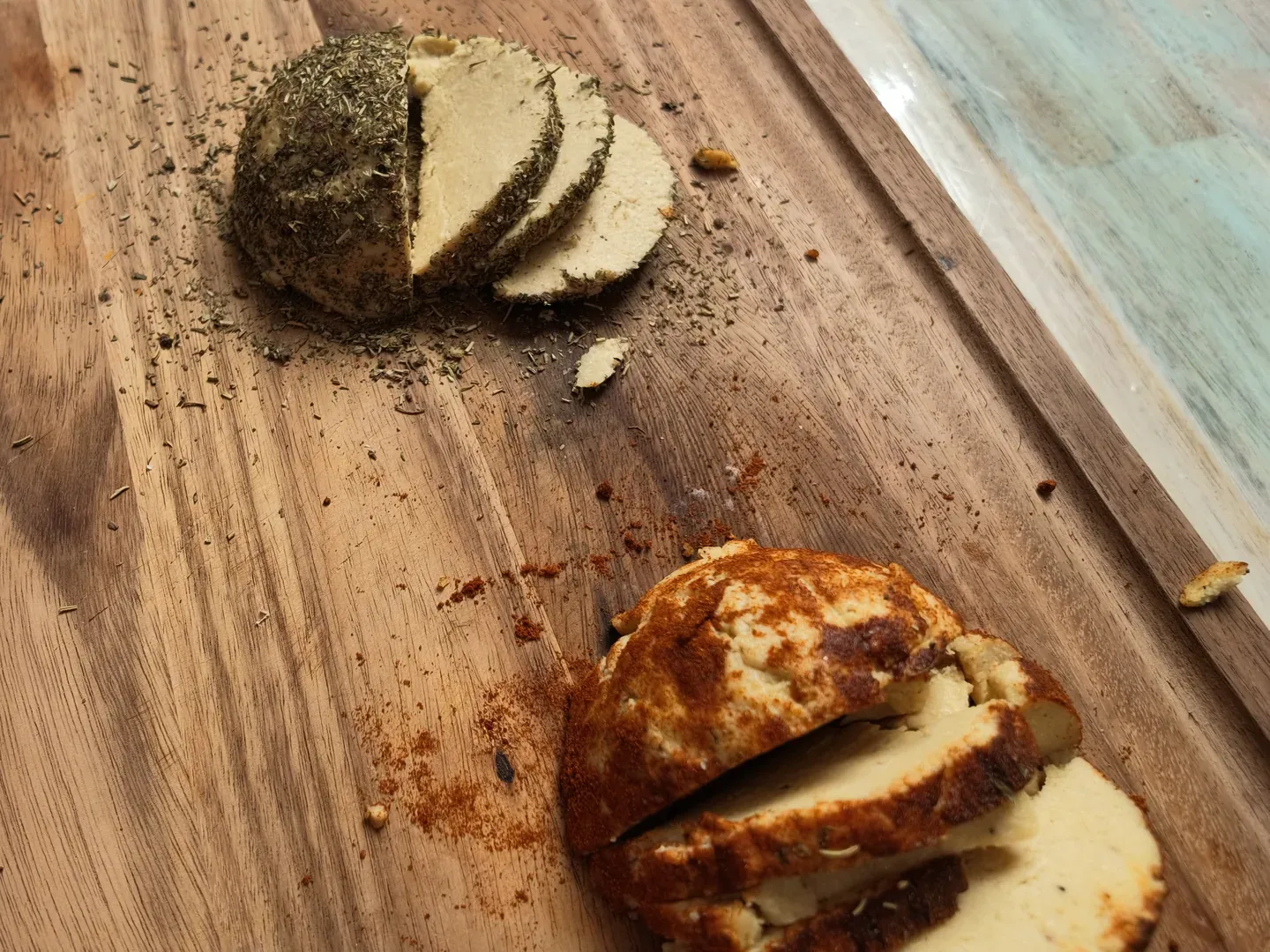 Two sliced vegan caciottas on a wooden board, one coated in dried oregano and one in red paprika, with spilled spices around them