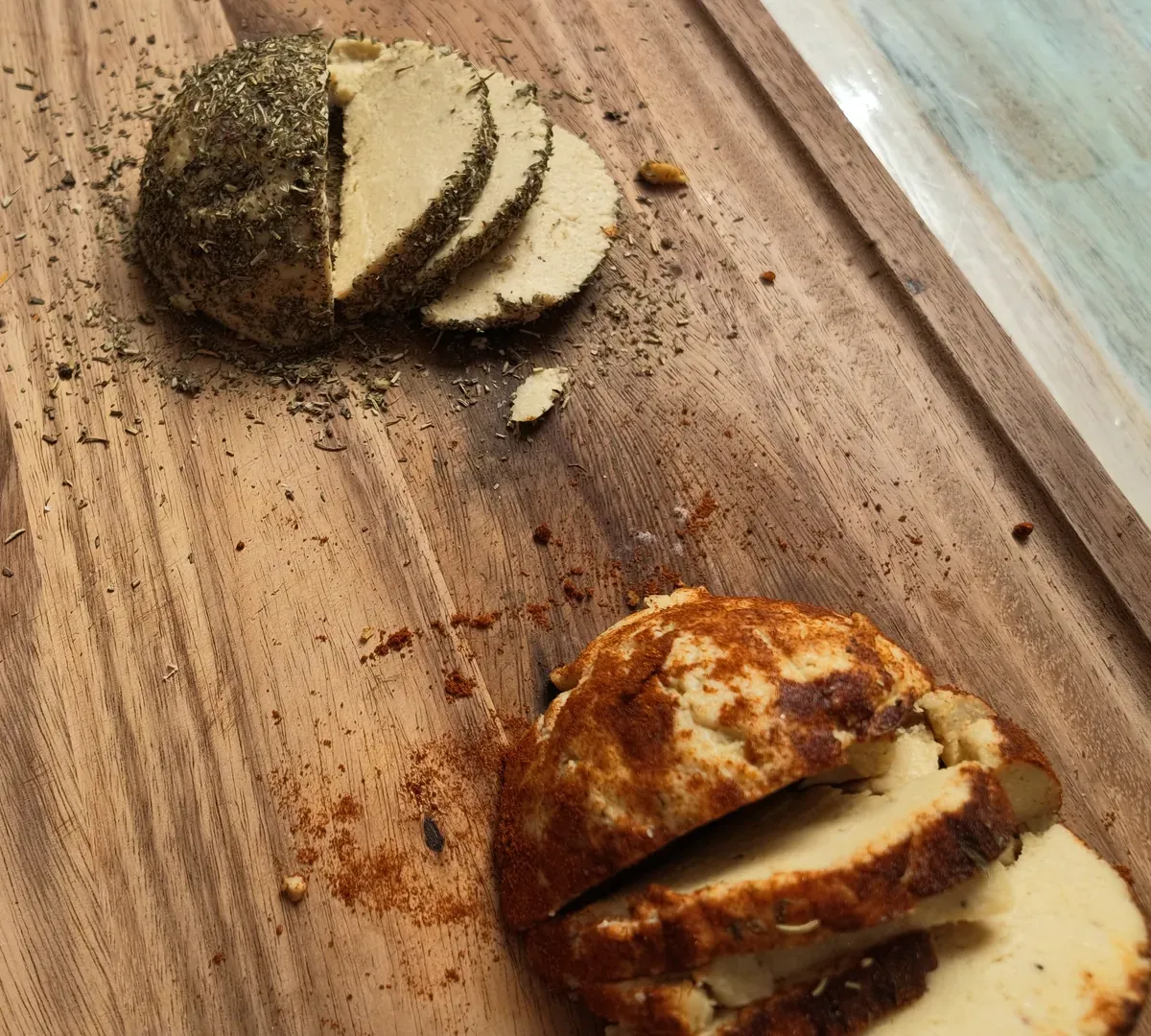 Due caciotte vegane affettate su un tagliere di legno, una ricoperta di origano secco e una di paprica rossa, con le spezie sparse attorno
