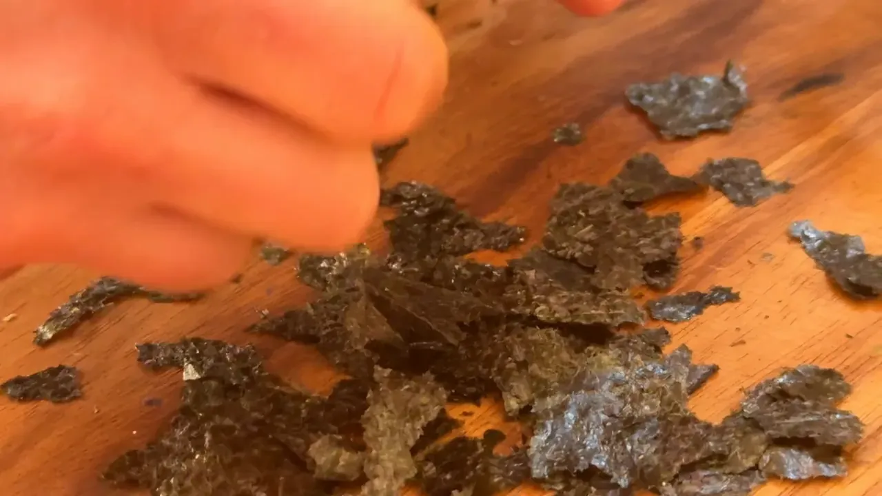 Fingers tearing dark nori sheets into small flakes on a wooden board