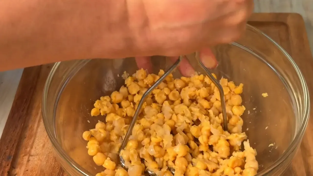 Chickpeas being coarsely mashed with a potato masher in a glass bowl