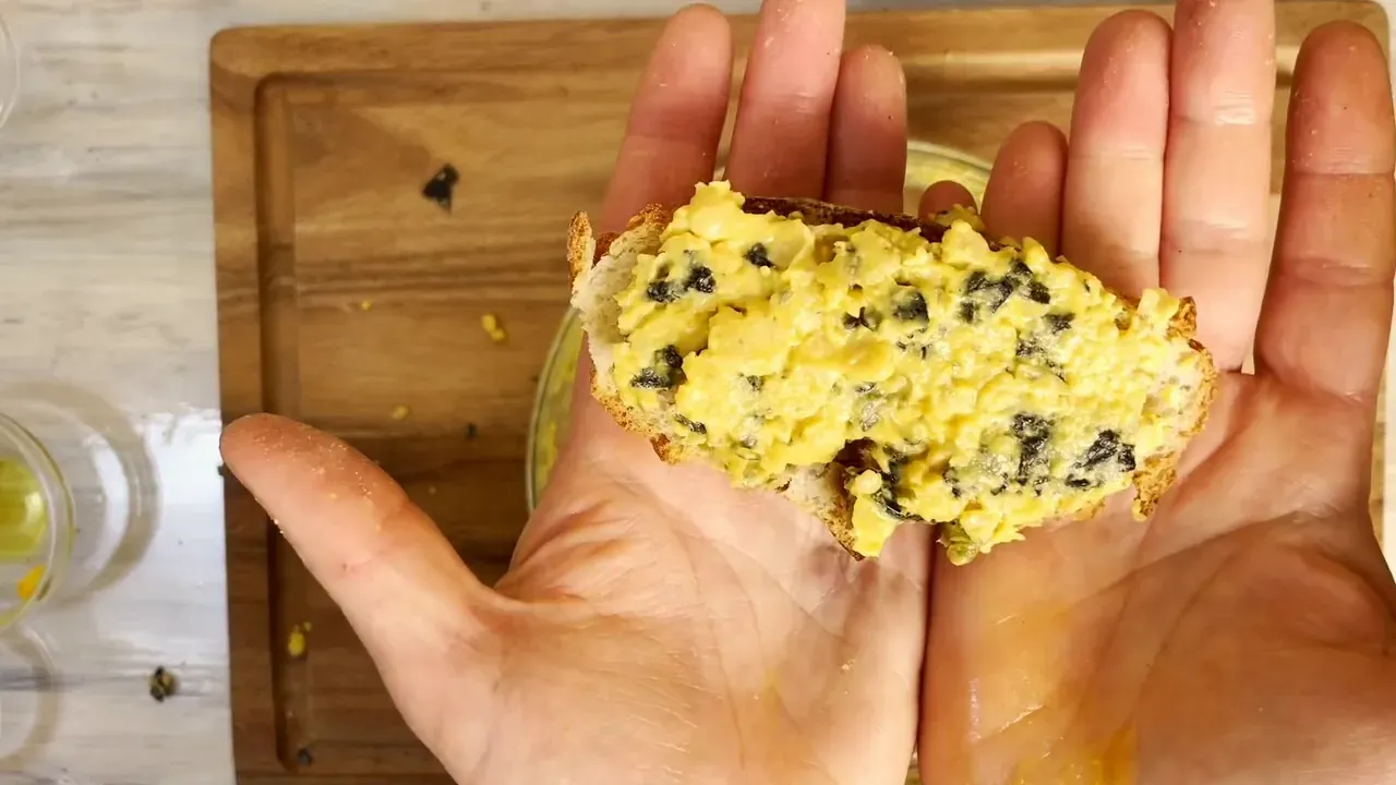 A piece of bread topped with yellow chickpea tuna speckled with nori, held in two open hands
