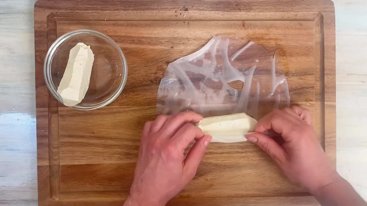 Immergo un foglio di carta di riso nel brodo caldo per circa 10 secondi, finché è flessibile ma non sfatto. Lo stendo sul piano di lavoro, dispongo metà del tofu al centro e lo arrotolo stretto a forma di salsiccia ripiegando le estremità.