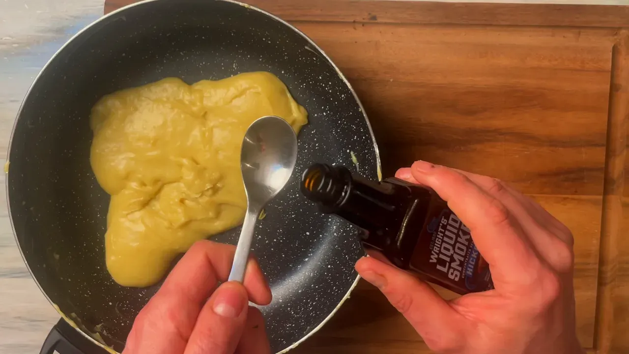 A bottle of liquid smoke tipped over a spoon above the cooked dough in the pan