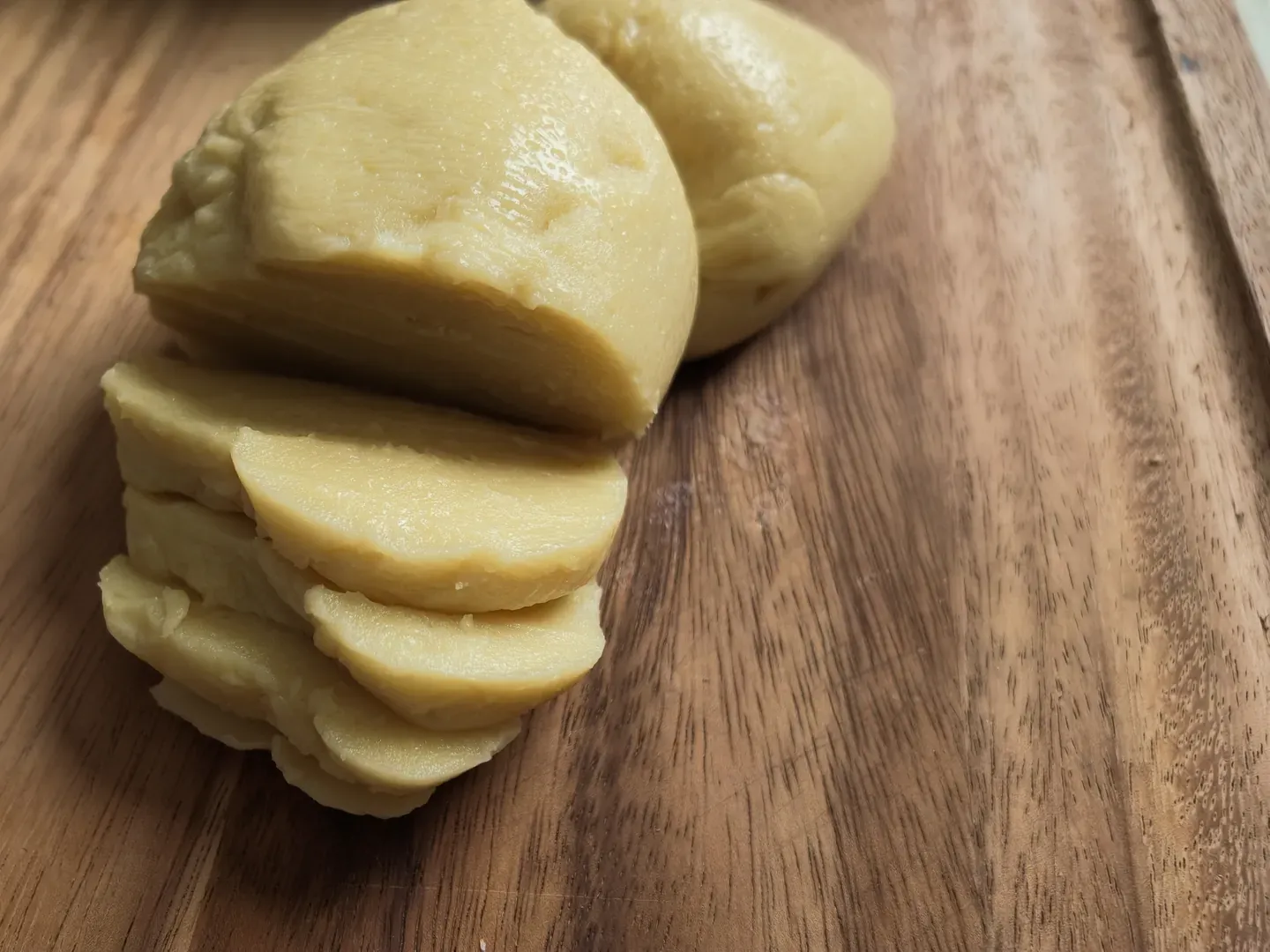 A pale yellow vegan provolone cut into thick slices on a wooden board, a second piece behind it