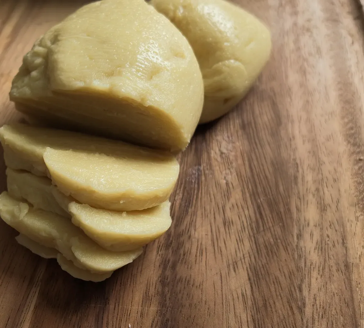 A pale yellow vegan provolone cut into thick slices on a wooden board, a second piece behind it
