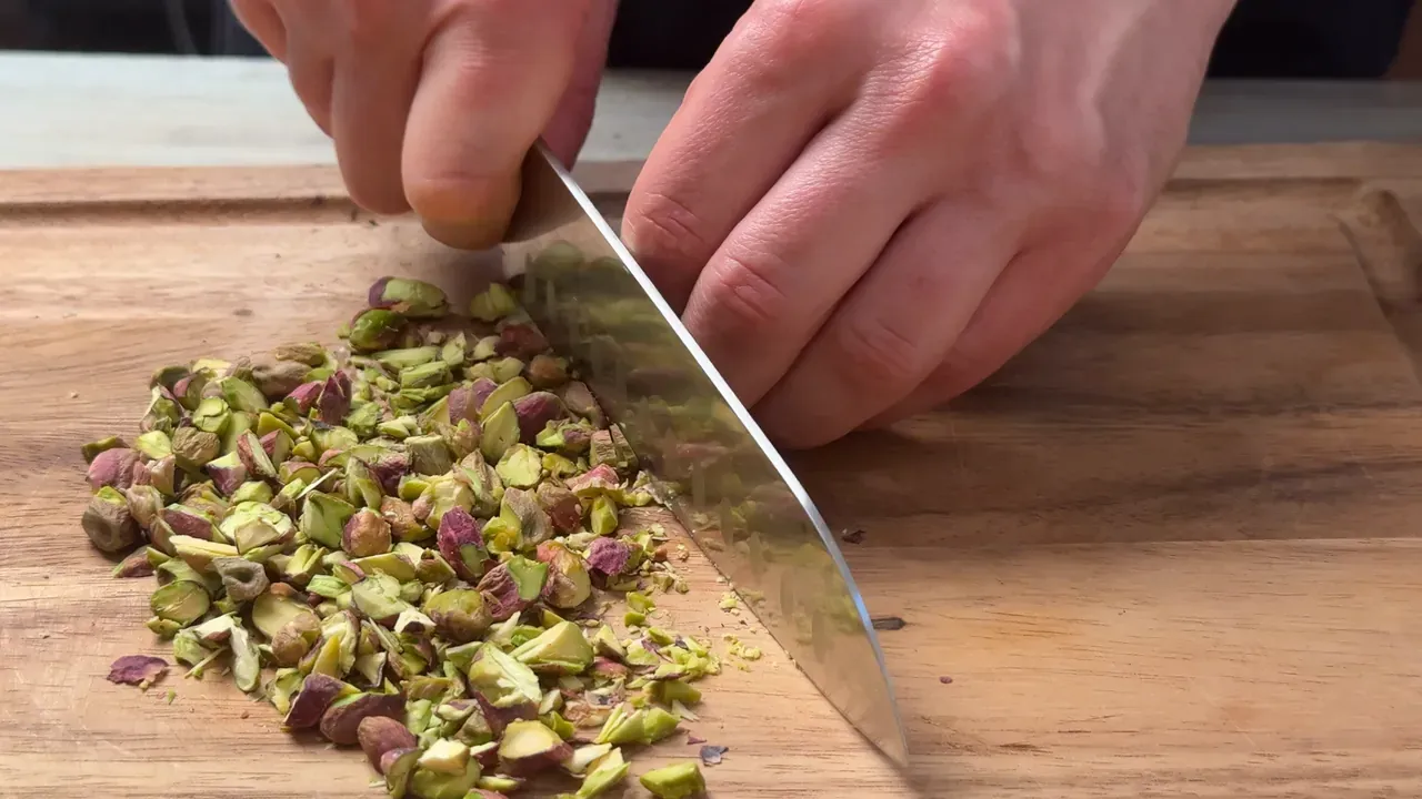 A chef's knife finely chopping shelled pistachios on a wooden board