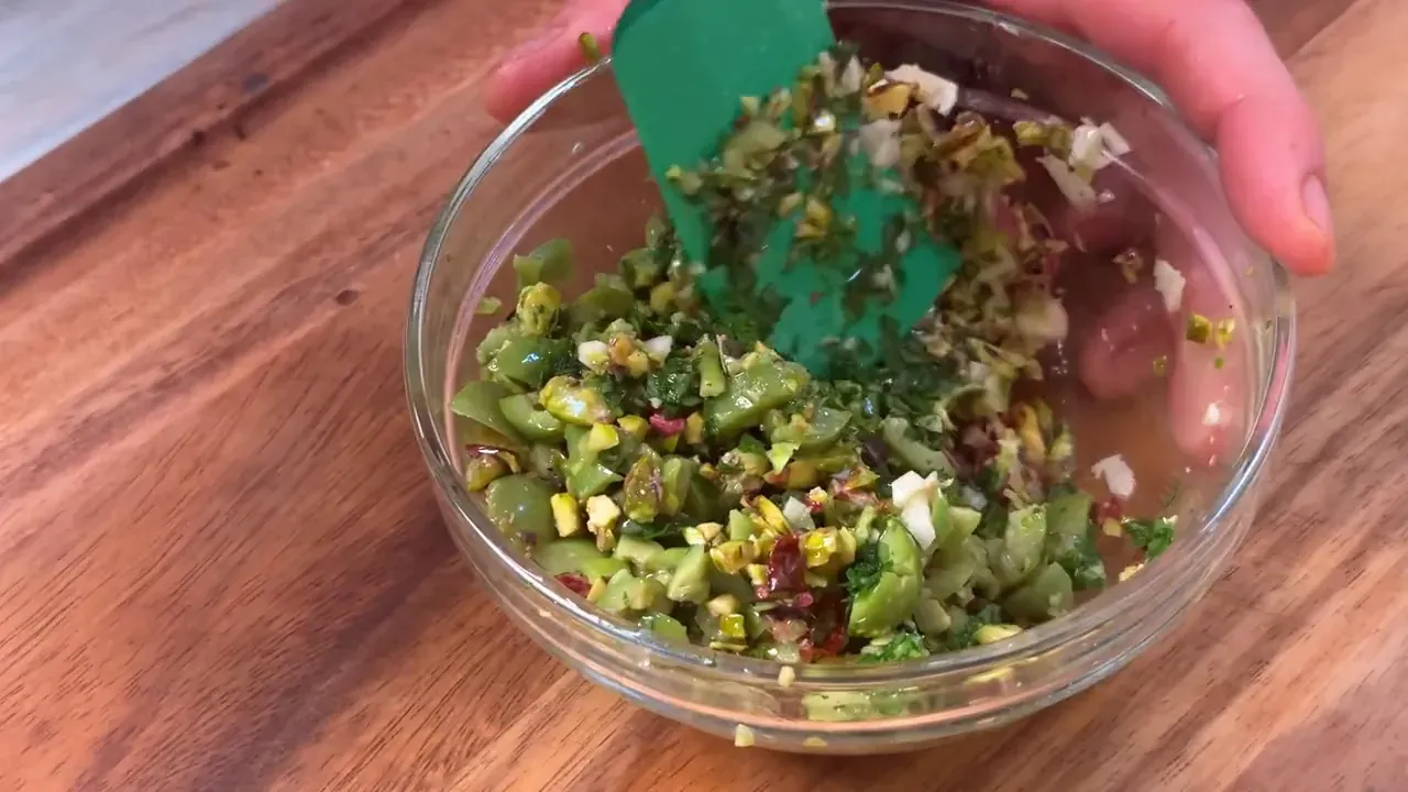 A green spatula mixing chopped olives, pistachios, parsley and chilli in a glass bowl