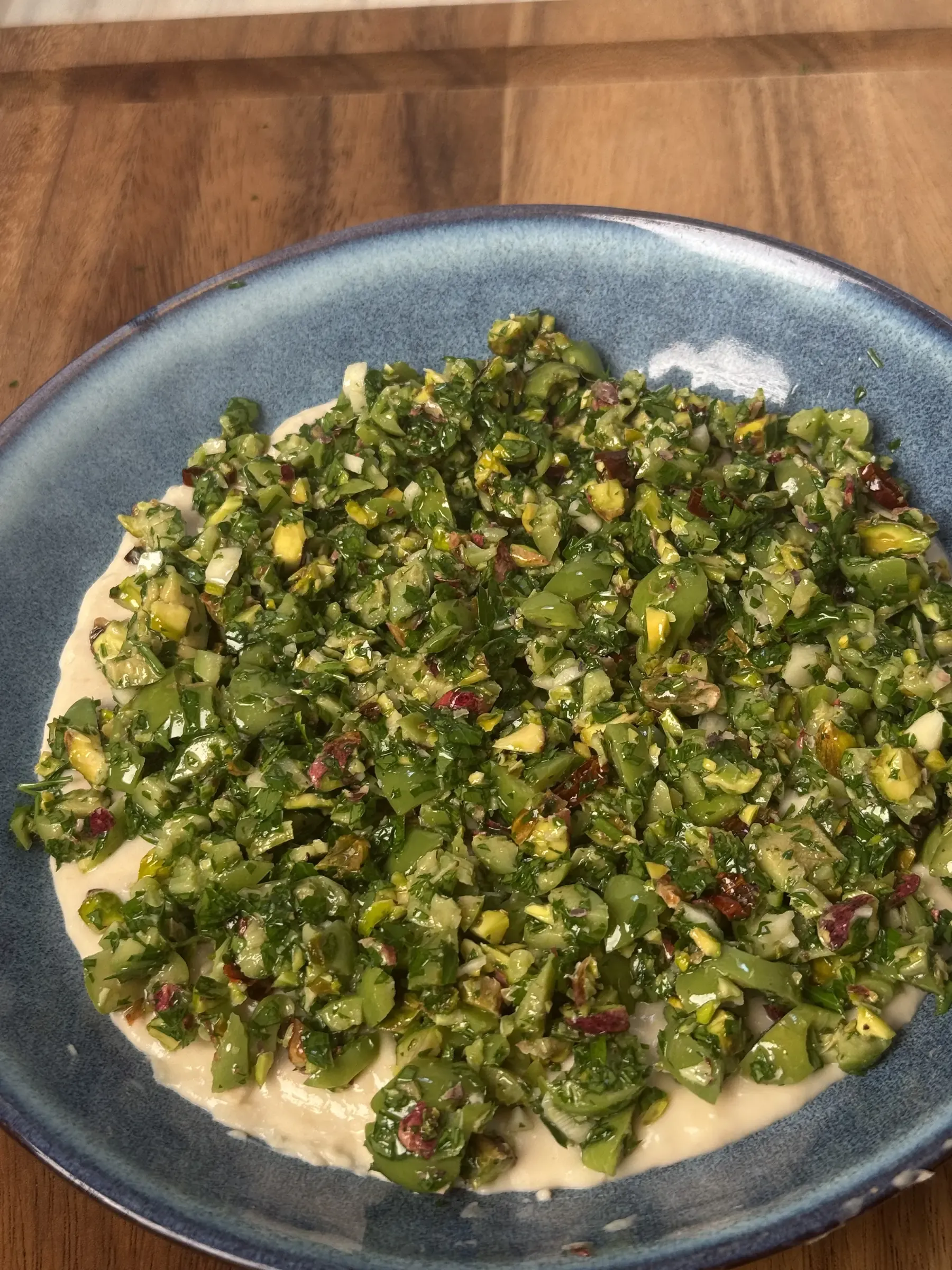 Blue ceramic plate of white bean cream covered with a chunky green salsa of chopped olives, pistachios, parsley and chilli, on a wooden board